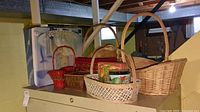 View of six assorted baskets in different sizes and colors including wicker and painted ones, two tins, and candle garden box on top of a cabinet.