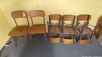 Eight children's desk chairs arranged against a wall: six smaller chairs with wooden seats and backs, metal legs; two larger chairs with attached wooden desks.