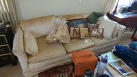 Beige three-seat upholstered couch with multiple decorative pillows and extra seat covers bagged on the couch.