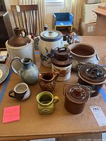 Overview photo of various stoneware and ceramic crocks, pitchers, and planters on a table in indoor lighting