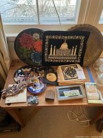 Overview of lot items on wooden table showing decorative brass plate, floral ceramic plate, Italian cotton squares, and various small decorative wall art pieces.