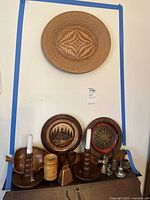 Photo showing large carved wood plate with woven pattern above smaller carved plates, brass figurines and wooden candlesticks on shelf.