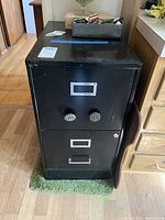 Full front view of the black metal two-drawer file cabinet showing drawer handles and label holders, placed on green mat.