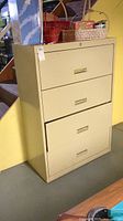 Front and side view of beige metal file cabinet with four drawers against yellow wall. Several woven baskets on top. Tag attached to cabinet.