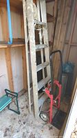 Full view of the aluminum folding ladder leaning against shelf with paint stains visible, along with red metal hand truck in front.