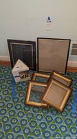 Six picture frames on patterned carpet floor against wall, including boxed Burnes triangular frame and five rectangular frames, three small gold-tone and two larger black bordered.
