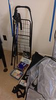 Overall view of black metal folding cart, walking sticks, boxed and packaged medical and hygiene items on floor near cart and bags