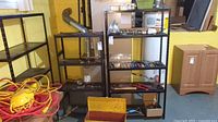Two black metal shelves with contents removed, showing wear and use marks, located in basement with yellow walls and other tools around.