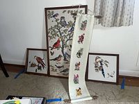 Four needlepoint artworks against wall showing detailed bird scenes, three framed and one scroll.