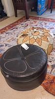 Two round ottomans placed on a patterned rug. Front ottoman is black faux leather with visible wear. Rear ottoman is larger, cushion style with faux animal hide and leather patchwork.