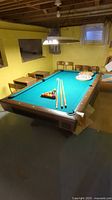 Pool table in basement with green felt, pool balls arranged in rack, two cues and bridge on table with yellow walls and small window in background.
