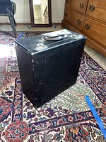 Closed black carrying case for the typewriter with a handle on top, sitting on a patterned carpet.
