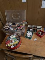 Photo of sewing notions showing clear plastic container filled with spools of thread, small boxes with buttons, and a round metal tin with sewing needles and accessories on a wooden table.