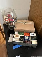 Large snifter glass filled with matchbooks, wooden cigar box placed next to it, additional matchbooks in a box in front.