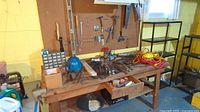 Wide view of wooden workbench with a variety of hand tools, extension cords, and a blue hard hat. Pegboard wall behind with hooks and additional tools.