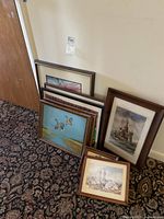 View of stacked framed artworks showing varied subject matter and frame styles on carpeted floor