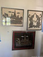 Three framed Asian themed needlepoint style wall art pieces, hung on a white wall. Two pieces show architectural and figure designs, one signed.