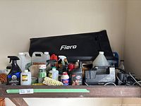 Various automotive cleaning liquids, sprays, brushes, red scrubbing pad, and Fiero branded black bag on wooden shelf.