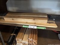 Shelf holding assorted wood pieces with various cuts and thicknesses, including flat planks and grooved slats, with metal mesh strip underneath