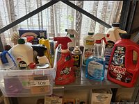 Photo shows a variety of household cleaning products on metal shelves, including recognizable brands Drano, Resolve, WD-40, Compliments, and Zep. Some bottles are spray bottles, others are pour bottles. Various levels of fullness are visible.
