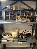 Two blue racks with multiple small drawers filled with assorted screws, nails, nuts, bolts, and miscellaneous hardware. Two containers with additional small items, plus a black battery charger and power bar on a metal shelf.