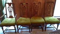 Four pecan wood dining chairs in a row showing detailed view of the front, highlighting turned spindle legs, carved backrests, and olive green cushioned seats.