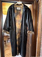 Front view of long black leather jacket hanging, showing cuff rolled with patterned lining visible, large front pockets and button fasteners.