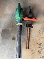 Overhead view of green electric leaf blower/vac with black blower tube alongside orange and black electric hedge trimmer with orange blade guard.