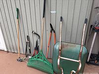 Full assortment of gardening tools including rake, clippers, cutters, weed hoe, seed spreader, and wheelbarrow leaning against shed.