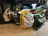 Five bags of assorted lawn and garden supplies placed on garage floor near shelf including Builder Max Grass Seed and Mushroom Manure fertilizer.