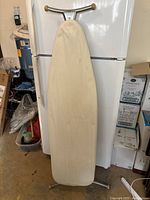Vintage ironing board with beige fabric cover and metal frame, standing upright against refrigerator.