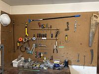 Wide view of pegboard wall with various hand tools hanging, parts, screws, and containers on the workspace surface.