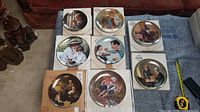 Eight Norman Rockwell painted plates displayed on a blanket, many shown with original boxes and certificates.