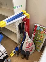Photo showing tennis rackets with Panther Series covers, cans of tennis balls, and badminton rackets leaning against a shelf along with folded soccer nets.