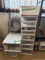 Wide view of the plastic storage tower with labeled sliding drawers alongside two large square plastic containers, showing assorted wiring and small parts inside.