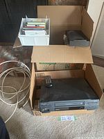 Overview of Toshiba VCR, remote, and cardboard boxes containing media items and cable cord.