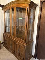 Front and side view of the vintage pecan china cabinet and hutch, highlighting the glass paneled upper section with shelves and lower wooden doors with metal handles.