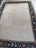 Full top view of stained beige area rug with dark floral border, showing marks and dirt.