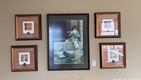 One photo showing four small square prints in gold-tone frames each with bronze coins mounted below the artwork, surrounding a larger black and wood toned framed print depicting a child feeding birds outdoors.