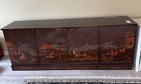 Front view of the large rectangular Asian lacquered chest showing hand-painted scenic panels depicting mountains, pagodas, boats, and figures.