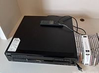 Black Sony five-disc CD player showing front panel with various function buttons, headphone jack, and disc selection buttons; includes remote control and several CDs stacked beside it.
