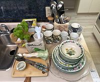 Overview of lot showing Royal Worcester floral china plates and bowls stacked, four matching mugs, metal cooking utensils in a container, small wooden cutting board with decorative garlic item and porcelain creamer, fresh basil plant, and kitchen disposable spoons.