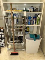 Wide view showing metal drying rack, broom, cleaning products on shelves, white trash bin, turquoise dustpan with brush, and paper towels.