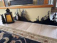 Metal wall art, metal bunny figurine, and candle lamp arranged on floor against a wall and window baseboard.