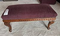 Side perspective of rectangular upholstered footstool showing burgundy fabric and carved wooden legs.