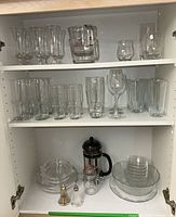 Full view of cupboard with three shelves containing a variety of clear glassware including goblets, tumblers, wine glasses, a measuring pitcher, glass plates, and kitchen items like salt and pepper shakers, plus a Bodum coffee press and a glass jar.