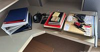 Photo showing a corner desk with multiple binders, notebooks, power adapters, and miscellaneous office supplies.