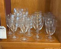 Eighteen clear glass stemware items arranged in rows inside wooden cabinet shelf.
