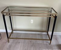 Straight-on photo showing full front view of rectangular glass hall table with metal frame and two glass shelves on light wood floor against cream wall.