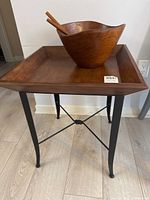Front view of wooden side table with square top holding wooden salad bowl with serving utensils.